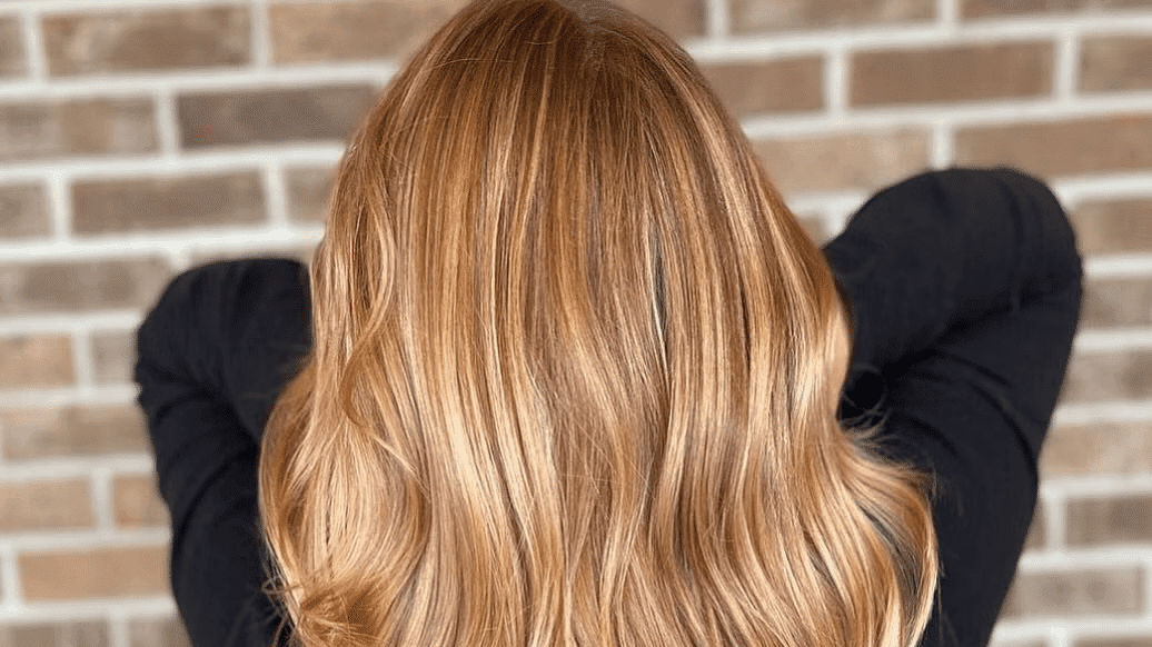 Wavy, golden blonde hair cascading down a person's back, set against a brick wall backdrop.